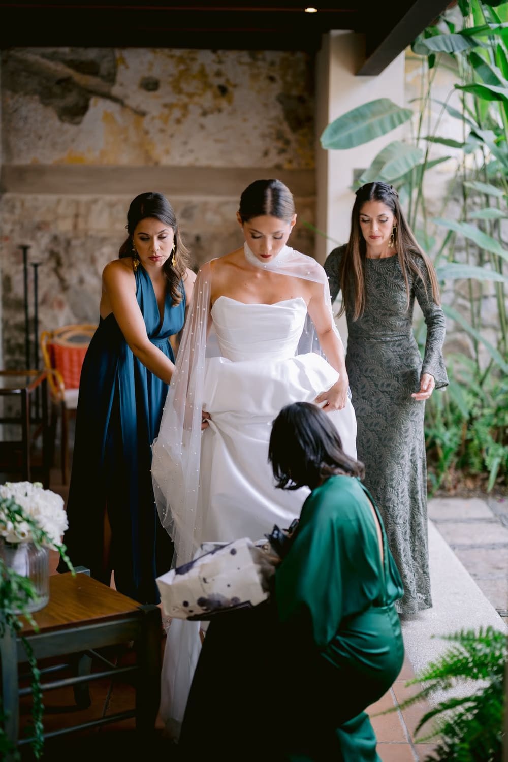 Bride stands flanked by her sisters as the seamstress kneels to adjust the gown — getting-ready moment with family in La Antigua.
