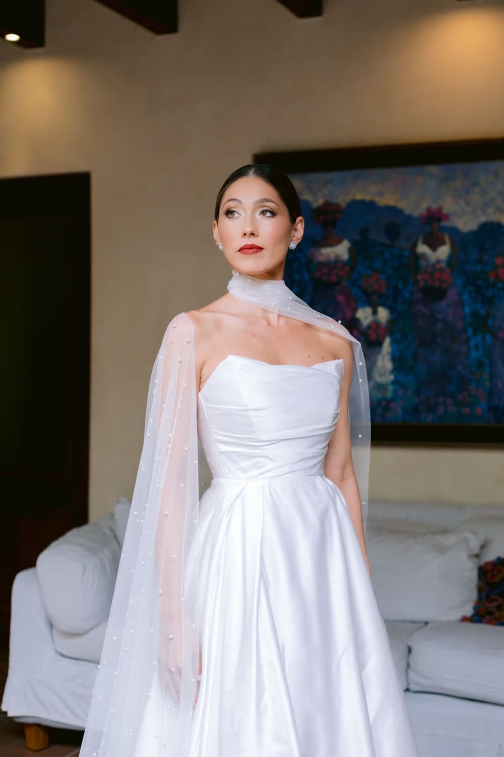 Bridal portrait inside the suite — satin gown and pearl-dotted cape glowing against warm walls and a vivid blue painting.
