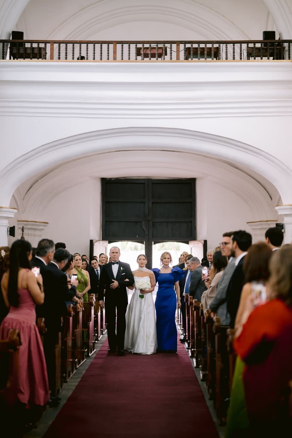 Bride walks down the aisle escorted by her parents as guests rise on either side — wedding processional in Antigua Guatemala.