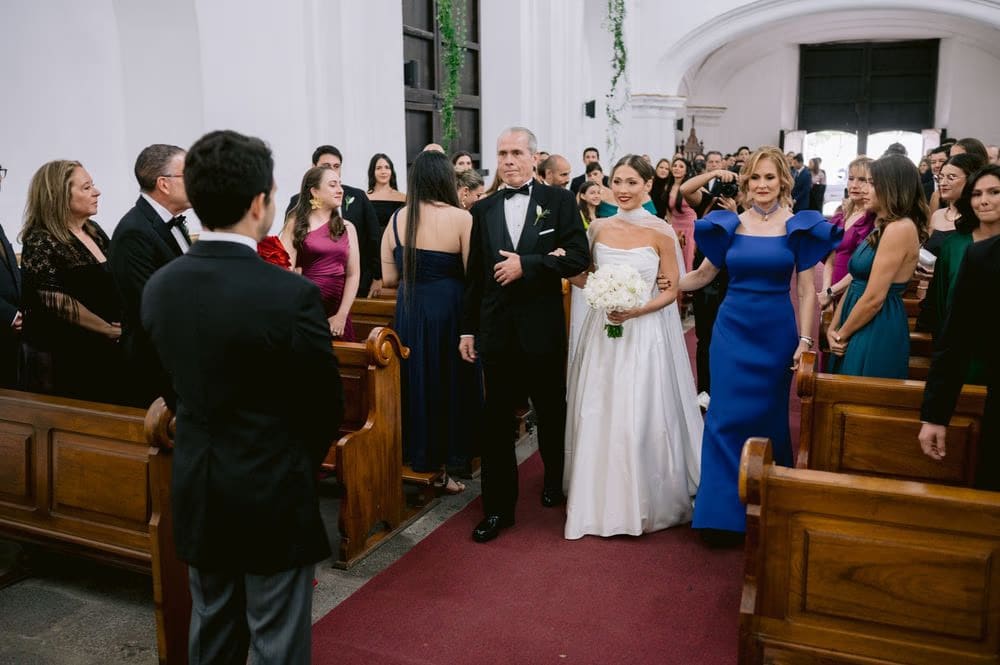 Bride walks the aisle between her father and mother as guests in colorful gowns turn to watch — family wedding processional.