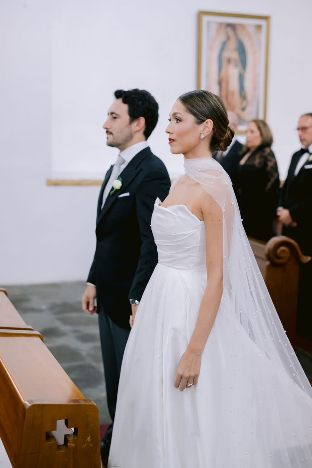 Bride and groom stand side by side before a Virgin of Guadalupe painting at the start of their wedding ceremony.