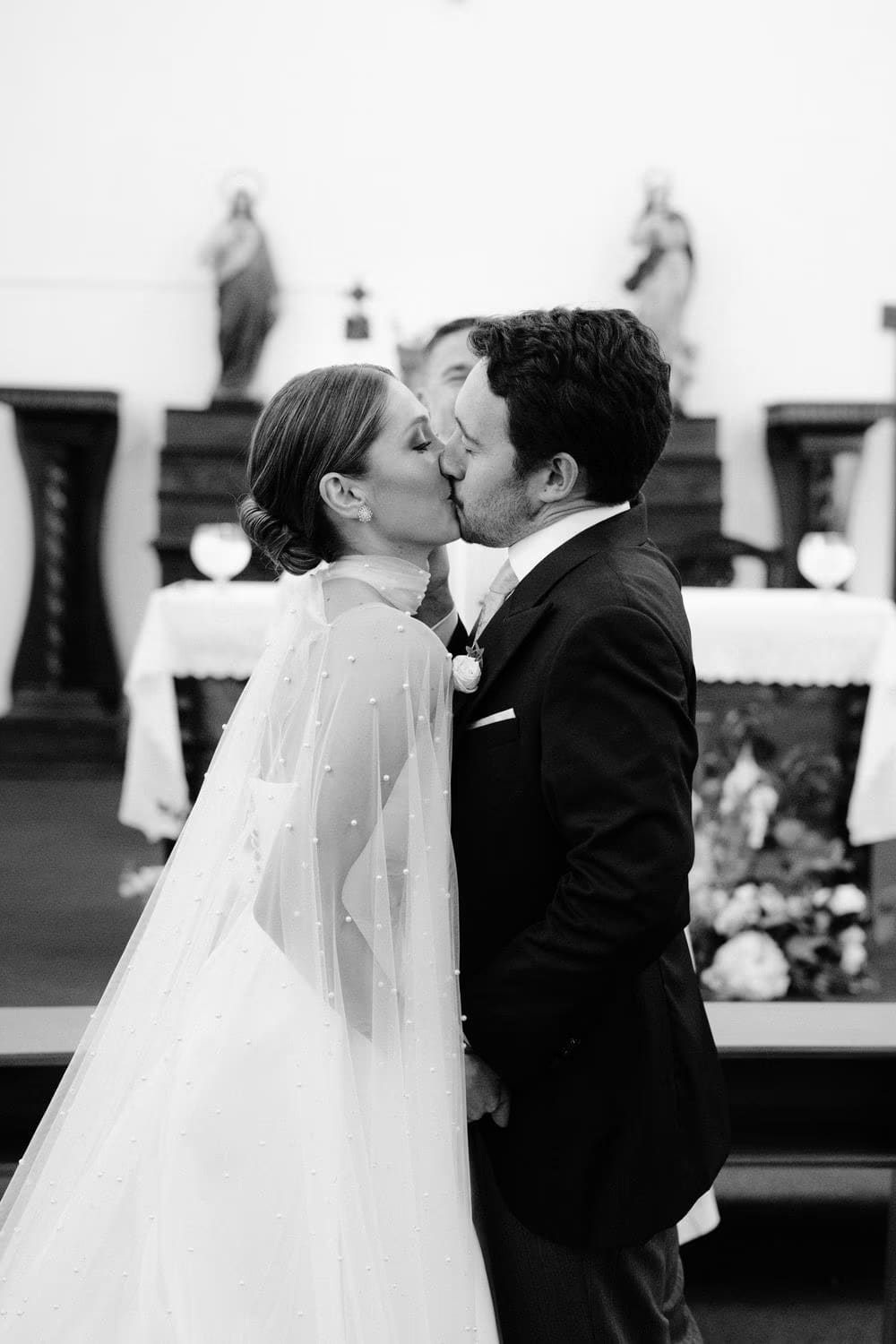 Andrea and Juan share their first kiss in black-and-white, framed by altar saints during their wedding ceremony in Antigua.