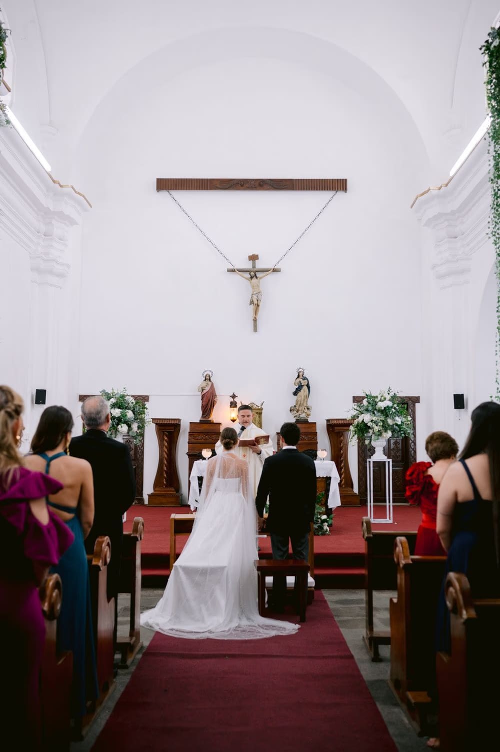 Wide aisle view of the wedding ceremony, priest reading at the altar between bride and groom — colonial church in Antigua.