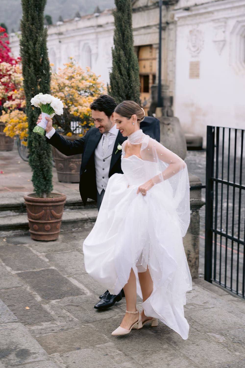 Groom raises the bouquet behind his bride as they cross a cobblestone plaza with yellow bougainvillea — joyful exit in Antigua.