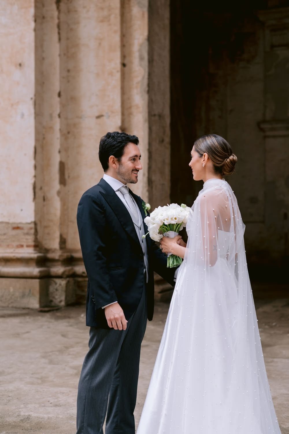 Couple smile at each other between weathered columns at Capuchinas Ruins, white bouquet between them — portrait in Antigua Guatemala.