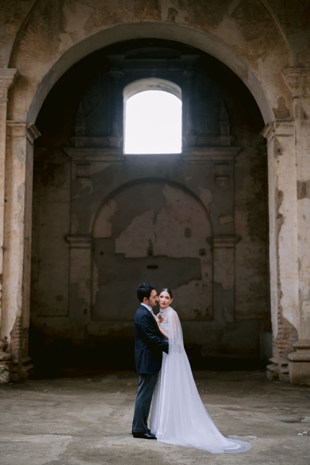 Couple framed beneath a tall arch and skylight inside Capuchinas Ruins — intimate destination wedding portrait in Antigua Guatemala.