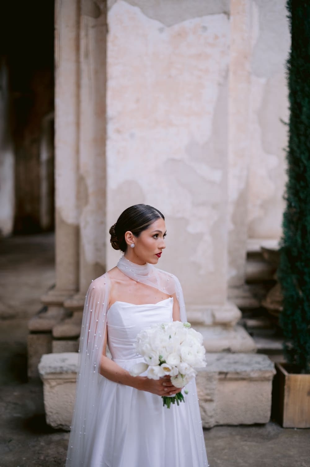 Bride stands holding her white bouquet beside a weathered cypress at Capuchinas Ruins — editorial portrait in colonial Antigua.