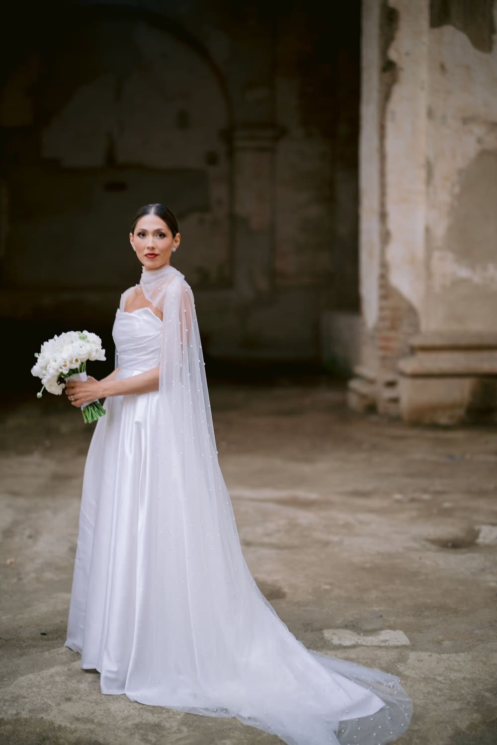 Full-length bridal portrait — pearl cape trailing behind, white bouquet in hand, weathered ruin walls behind in Antigua.