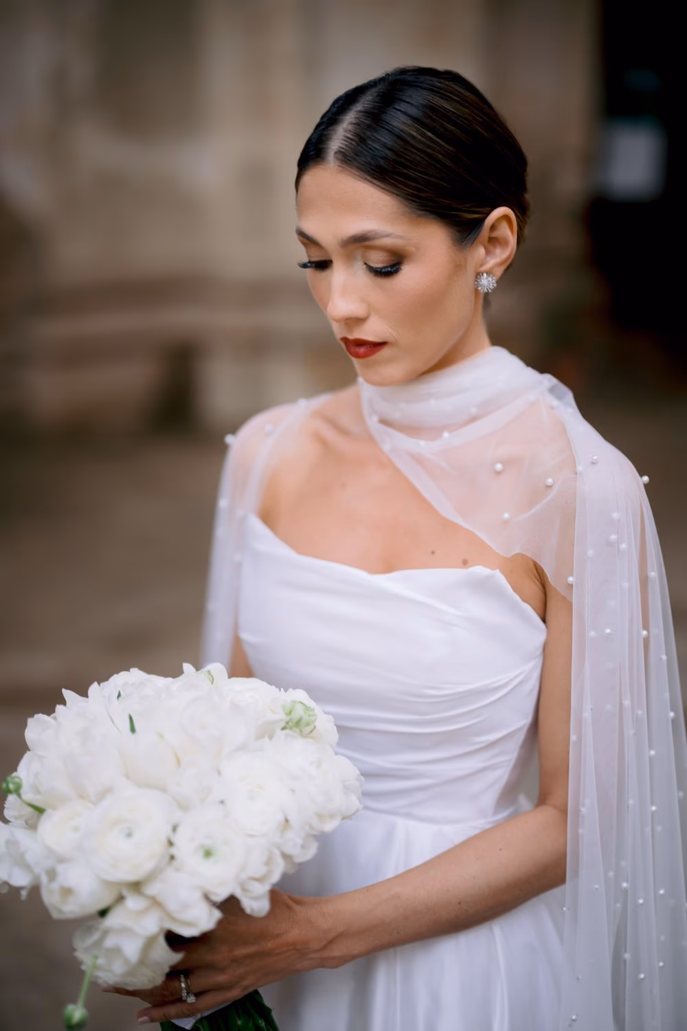 Close bridal portrait — bride looks down toward her white ranunculus bouquet, diamond rings catching the light.