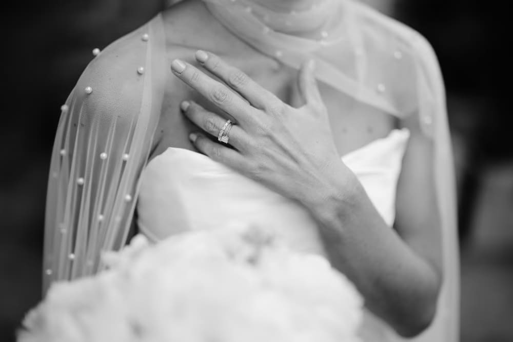 Black-and-white detail — bride's hand resting on her chest, wedding band visible through the sheer pearl cape.