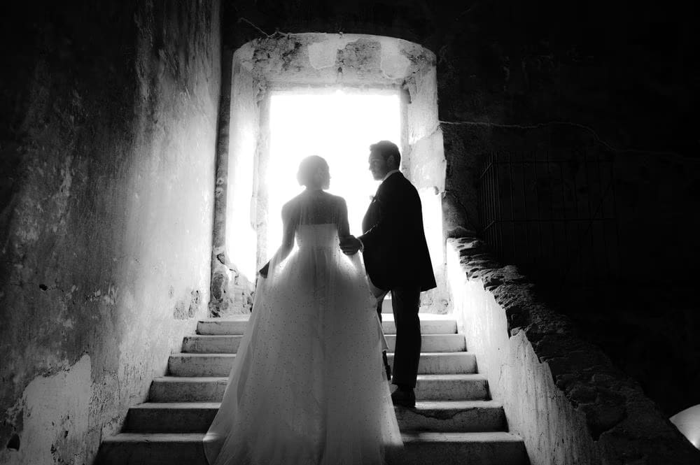 Silhouette of bride and groom holding hands on a stone staircase against bright daylight — black-and-white wedding moment.