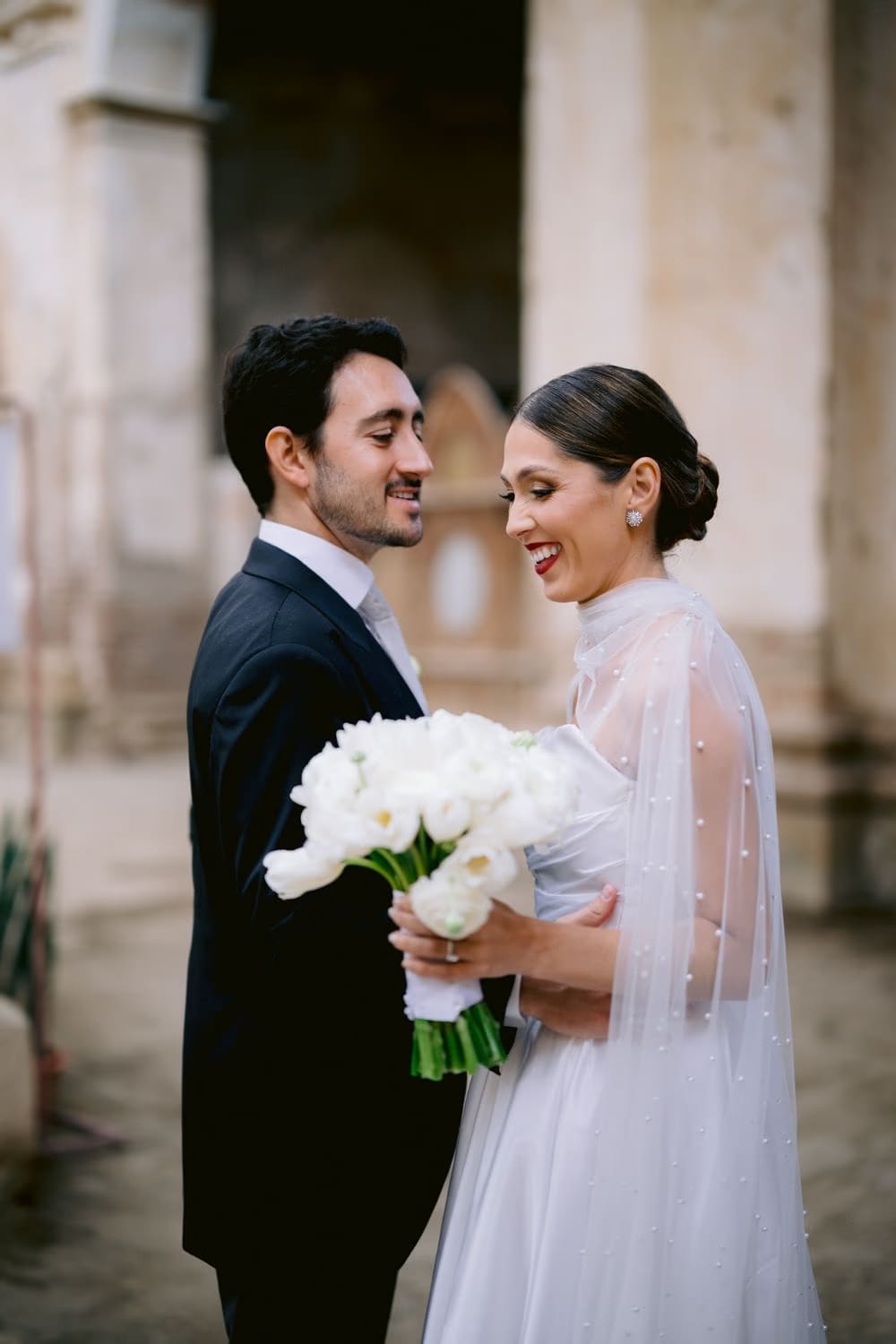 Andrea and Juan laugh together holding the white bouquet between them outside Capuchinas Ruins in Antigua Guatemala.