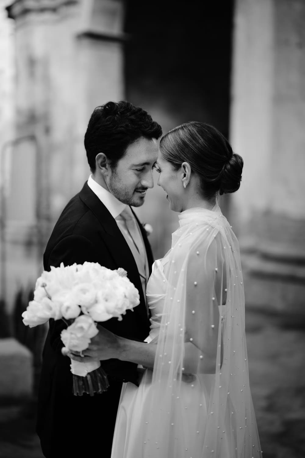 Black-and-white close portrait — bride and groom touch foreheads, white bouquet between them in colonial Antigua.