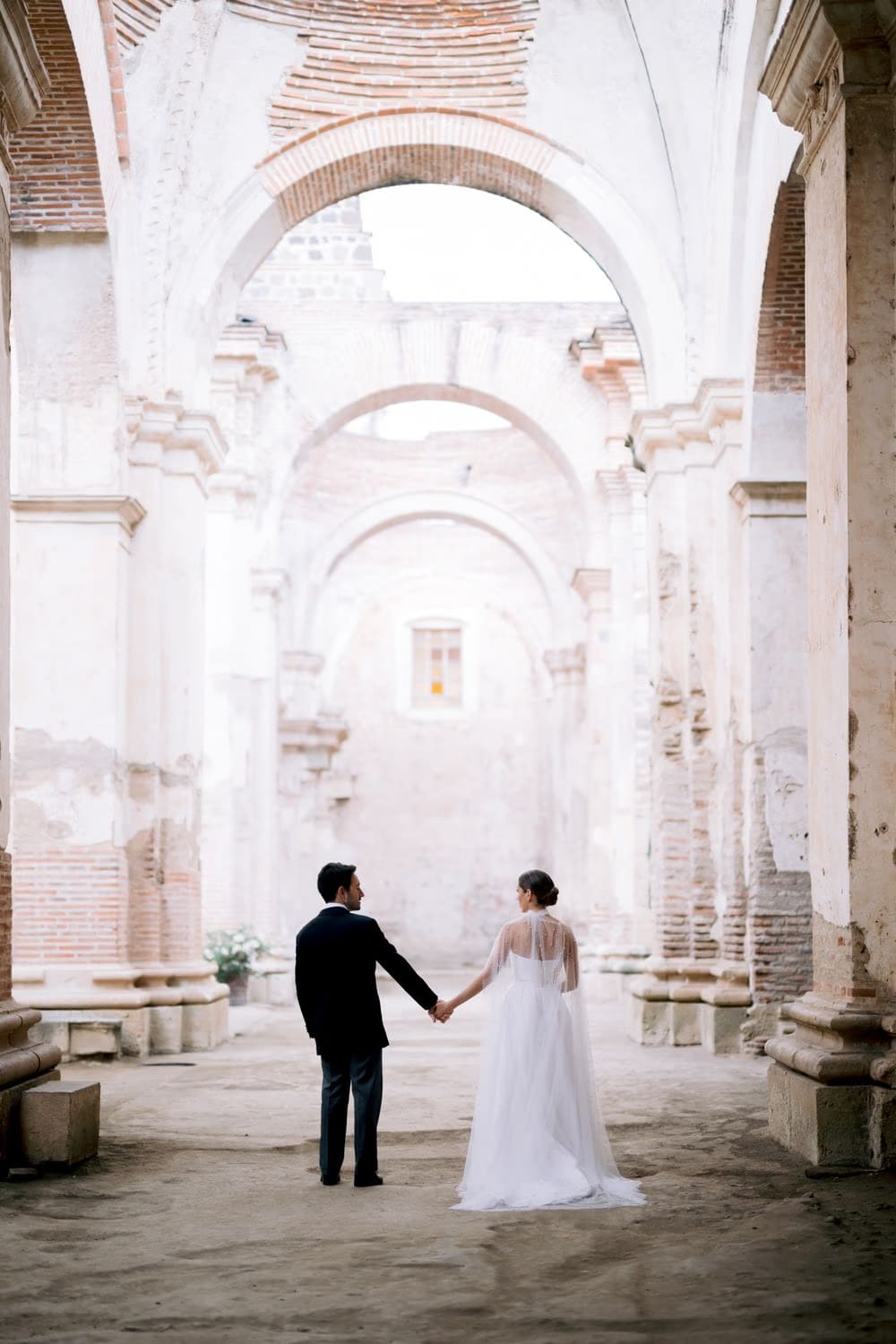 Couple stand hand-in-hand in the long arcade of brick arches at Capuchinas Ruins — destination wedding portrait in Antigua Guatemala.