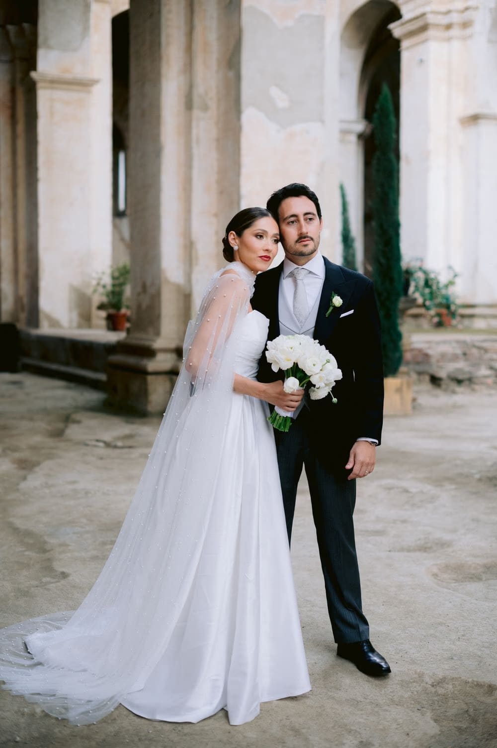Bride rests her cheek on the groom's shoulder in the cloister courtyard — editorial wedding portrait in colonial Antigua.