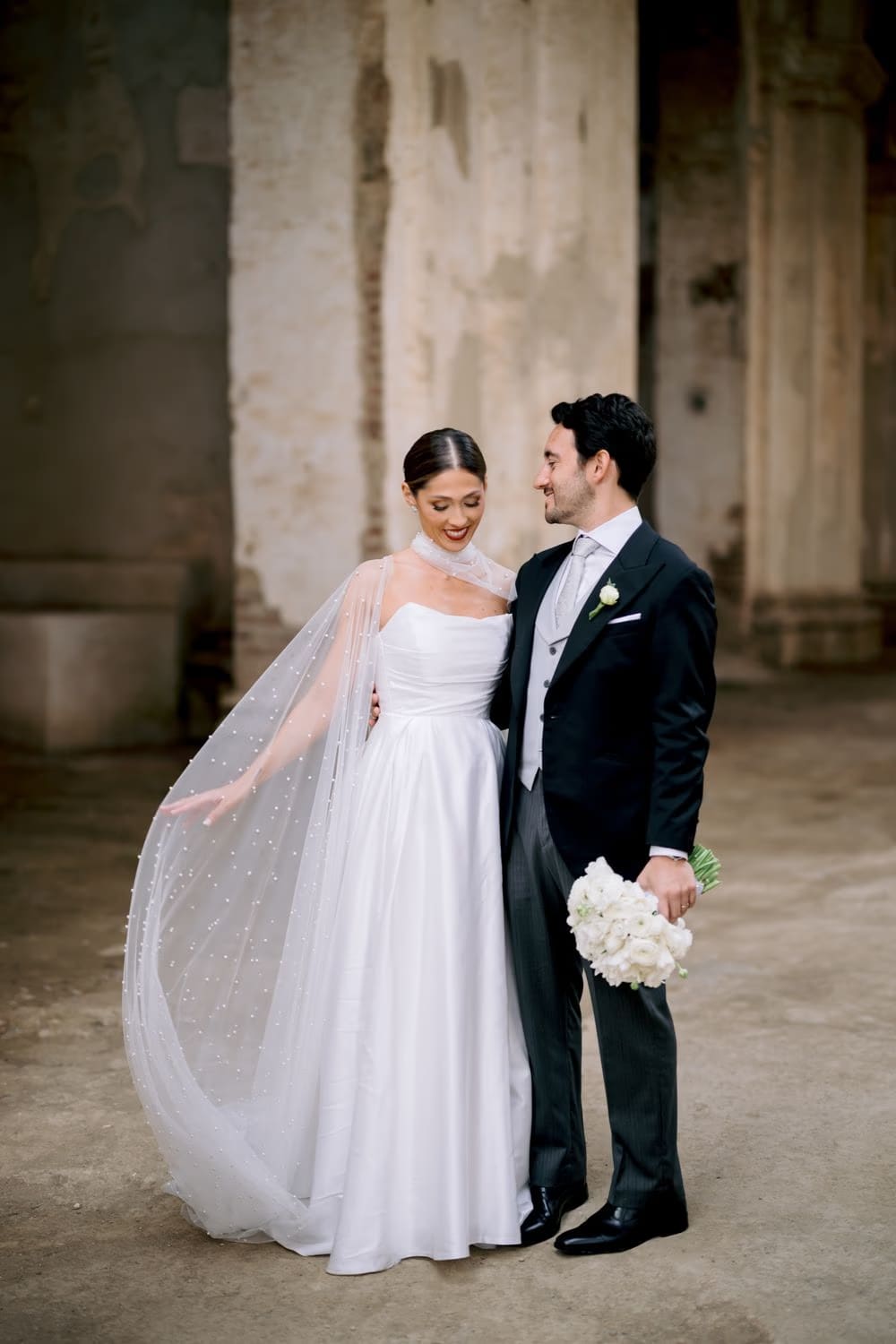 Pearl-dotted cape flutters between bride and groom as they laugh in the cloister at Capuchinas Ruins — Antigua Guatemala.