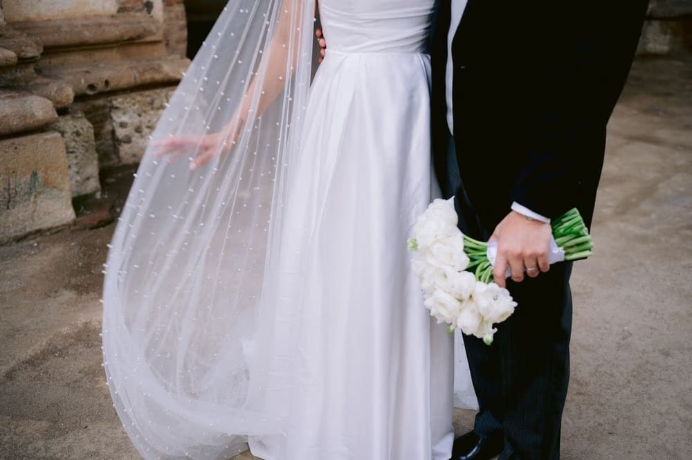 Cropped detail — pearl cape and white bouquet held between bride and groom at colonial wedding portrait.
