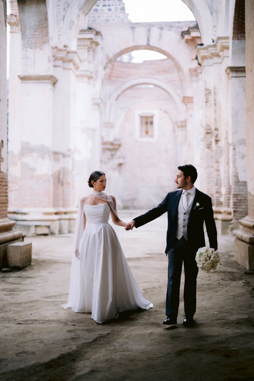 Bride and groom hold hands as they walk through the misty nave of Capuchinas Ruins — destination wedding in Antigua Guatemala.