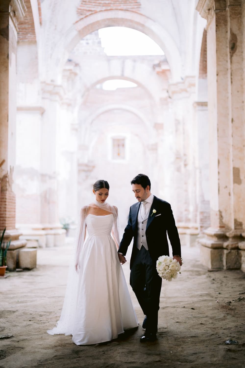 Bride and groom stroll hand-in-hand through soft light filling the long arcade of the ruined nave — La Antigua wedding portrait.
