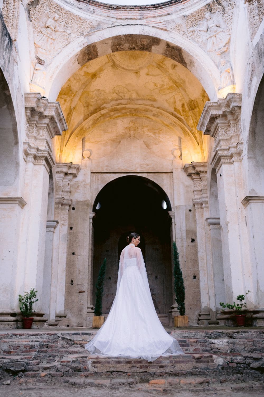 Bride stands alone with her back to the camera under the frescoed dome of Capuchinas Ruins, train flowing behind her.