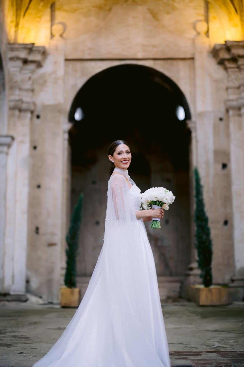 Bride smiles back over her shoulder holding her bouquet beneath an arched portal — colonial wedding portrait in Antigua.