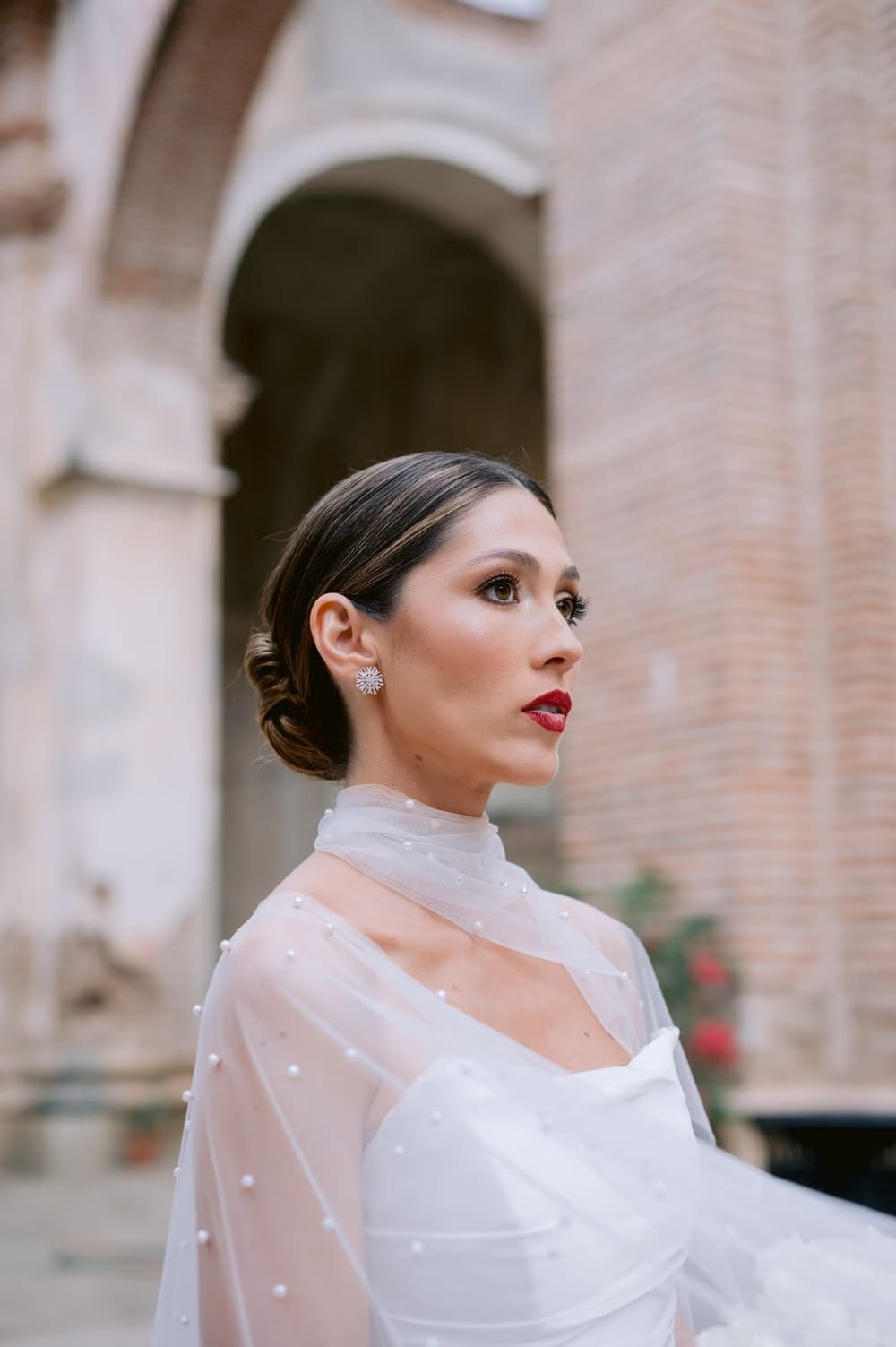 Profile bridal portrait against a soft pink brick wall — pearl cape and diamond earring detail.