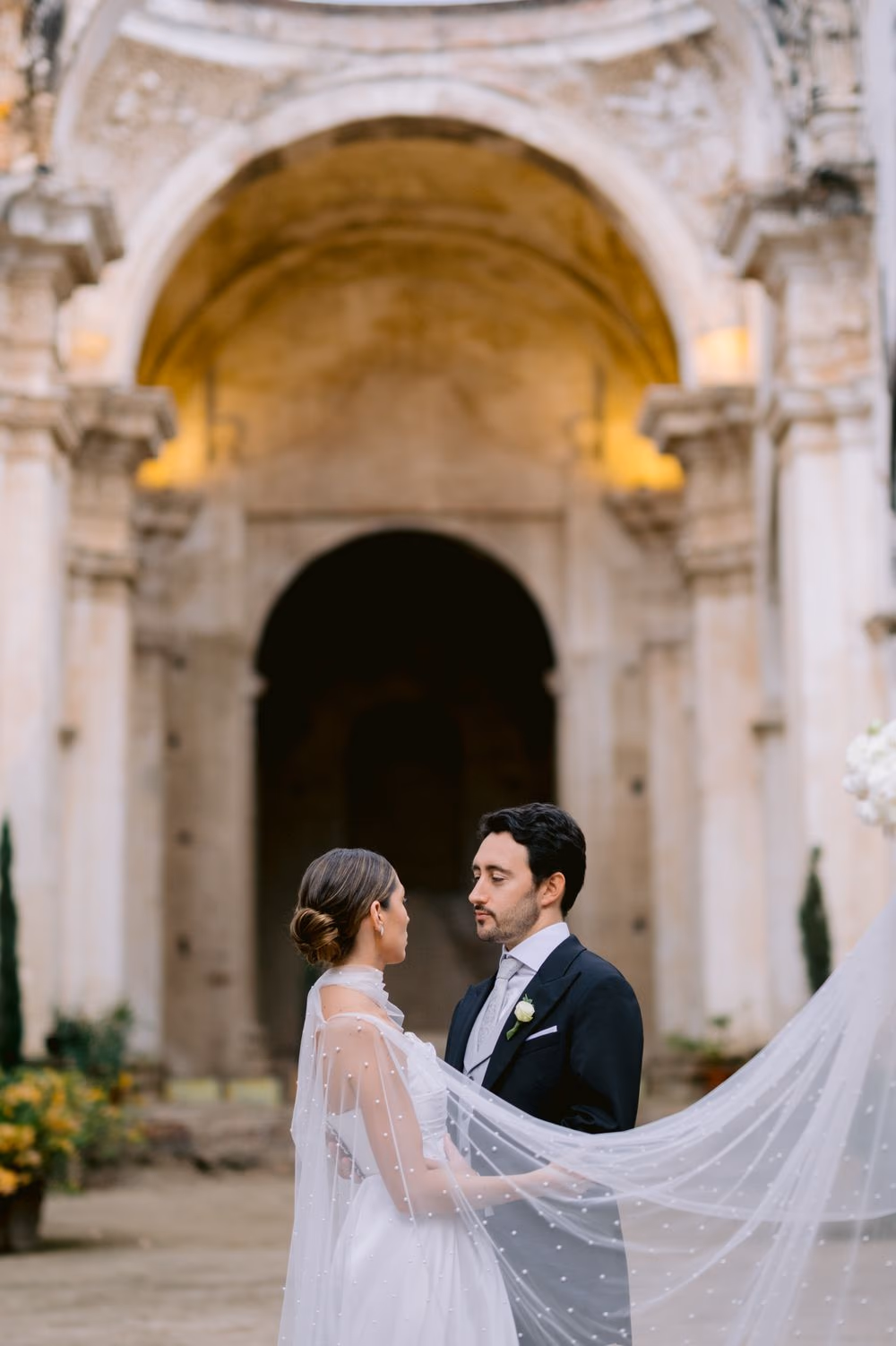 Bride and groom stand close as her veil cape sweeps across him beneath the arched portal of Capuchinas Ruins.