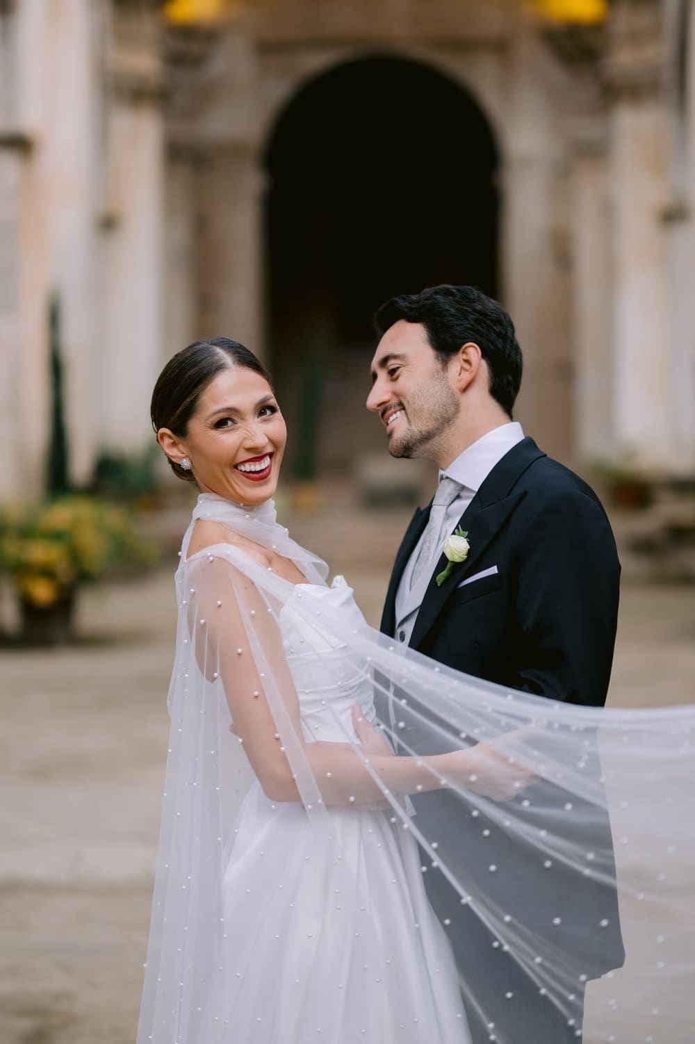 Couple laugh together in the cloister courtyard, the bride's cape catching the light around them in colonial Antigua.