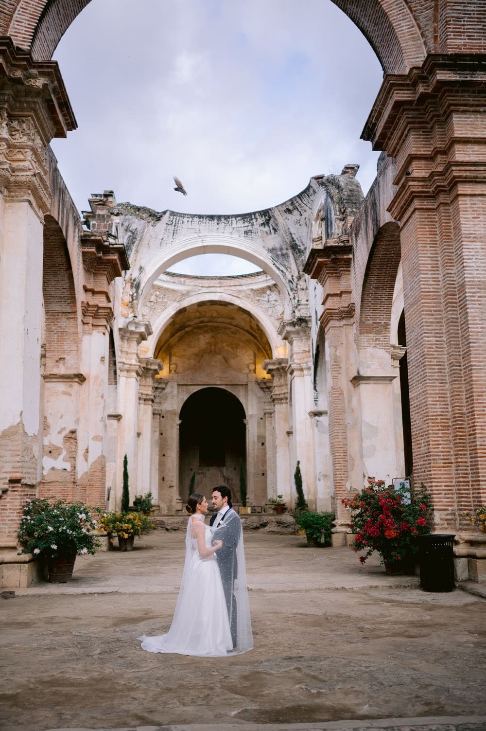 Wide view of the bride and groom under the open dome of Capuchinas Ruins, a bird passing across the sky overhead.