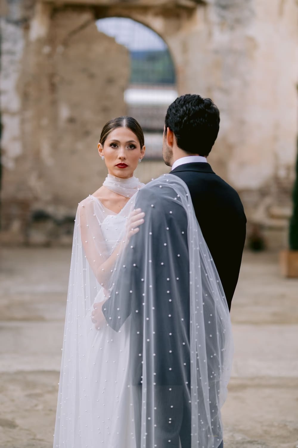 Bride glances at the camera over the groom's shoulder, his back turned — editorial wedding portrait at La Antigua ruins.