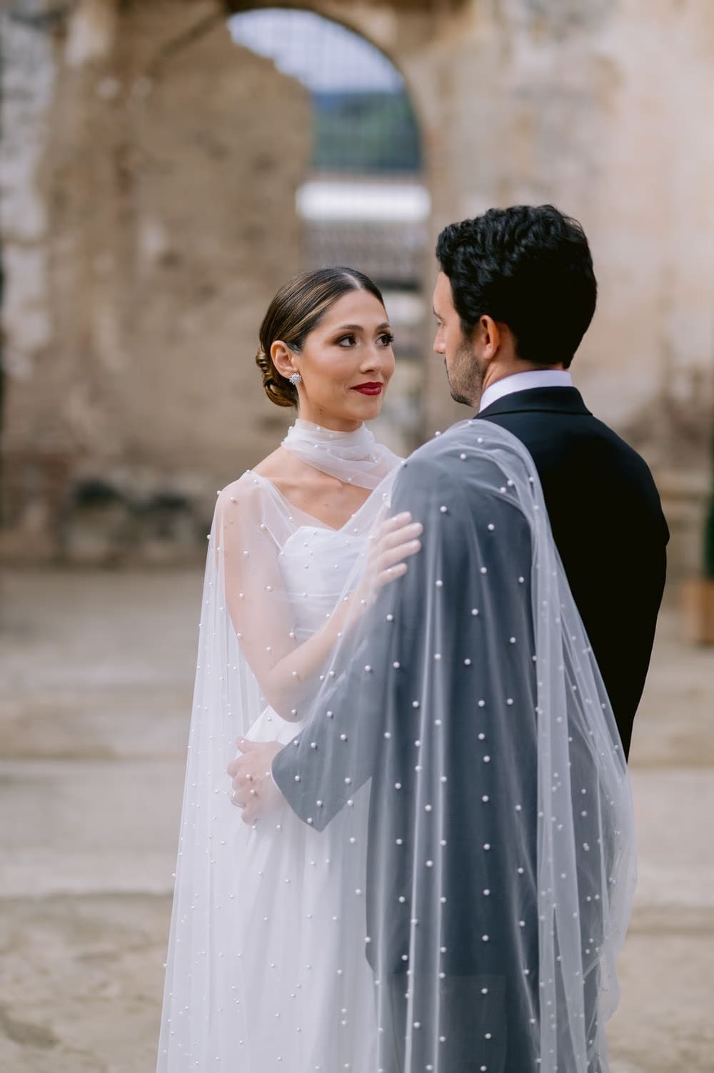 Couple face each other across a courtyard, the bride's hand resting on the groom's chest in a tender wedding portrait.