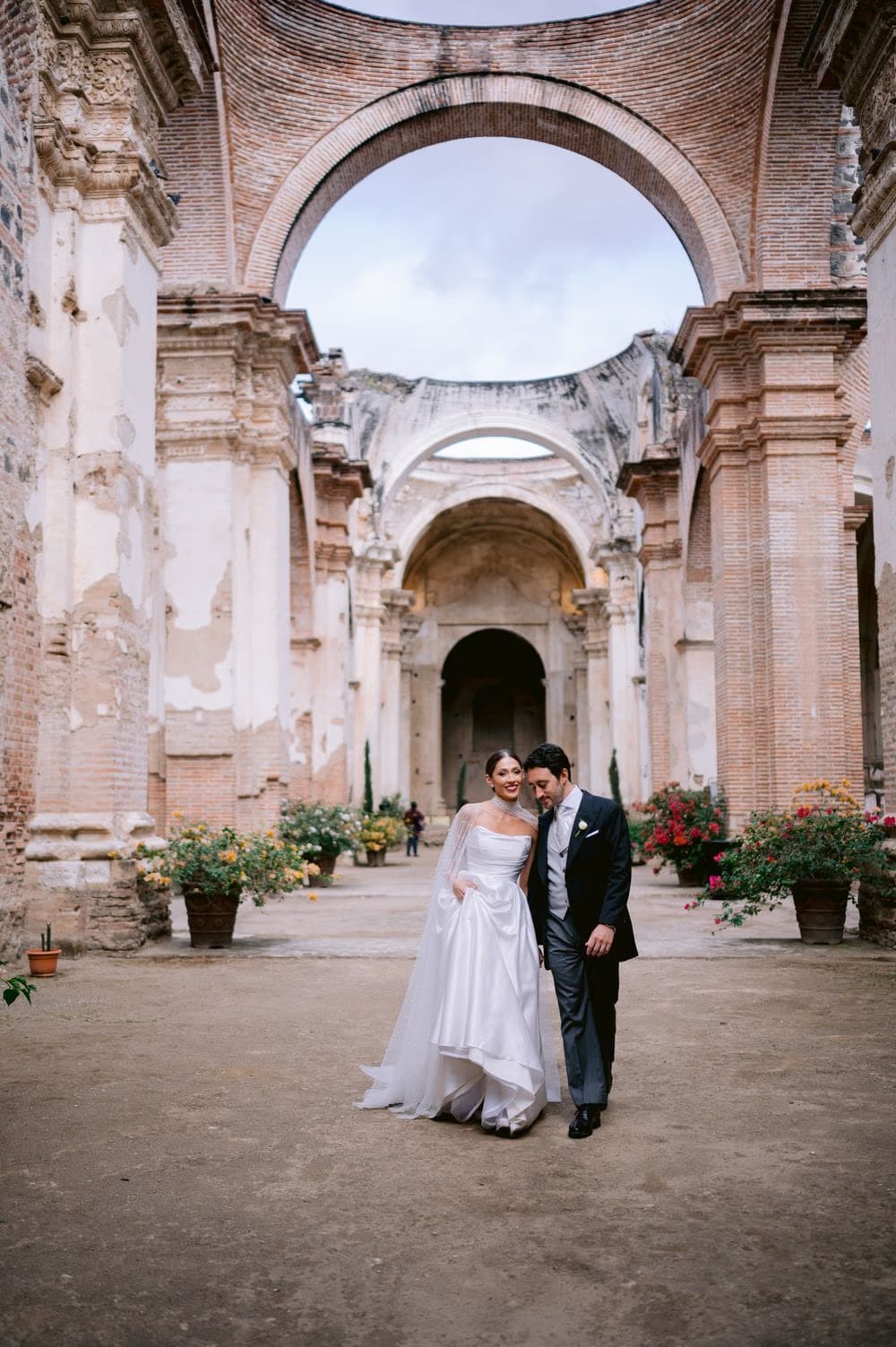 Couple stand smiling beneath the towering arches of Capuchinas Ruins, flowers blooming on each side — Antigua Guatemala wedding.