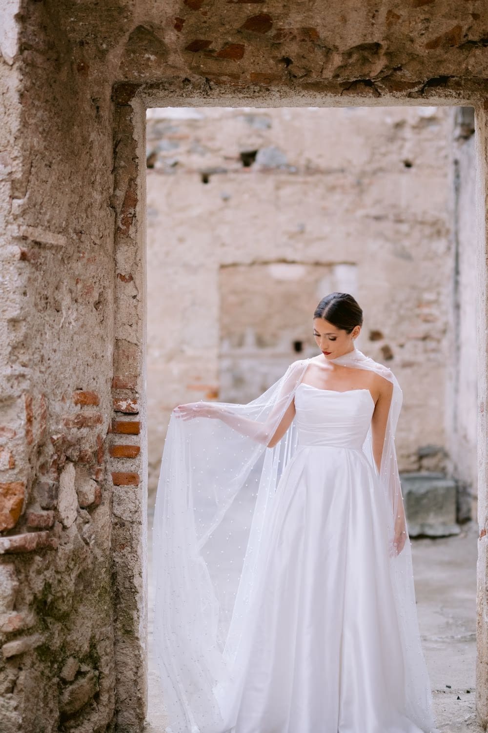 Bride lifts her cape with one hand inside a brick-walled doorway — ruin portrait at colonial Antigua wedding.
