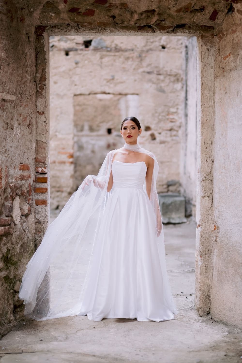 Bride centered under a rustic stone doorway with her tulle cape held outward — editorial wedding portrait at Capuchinas Ruins.