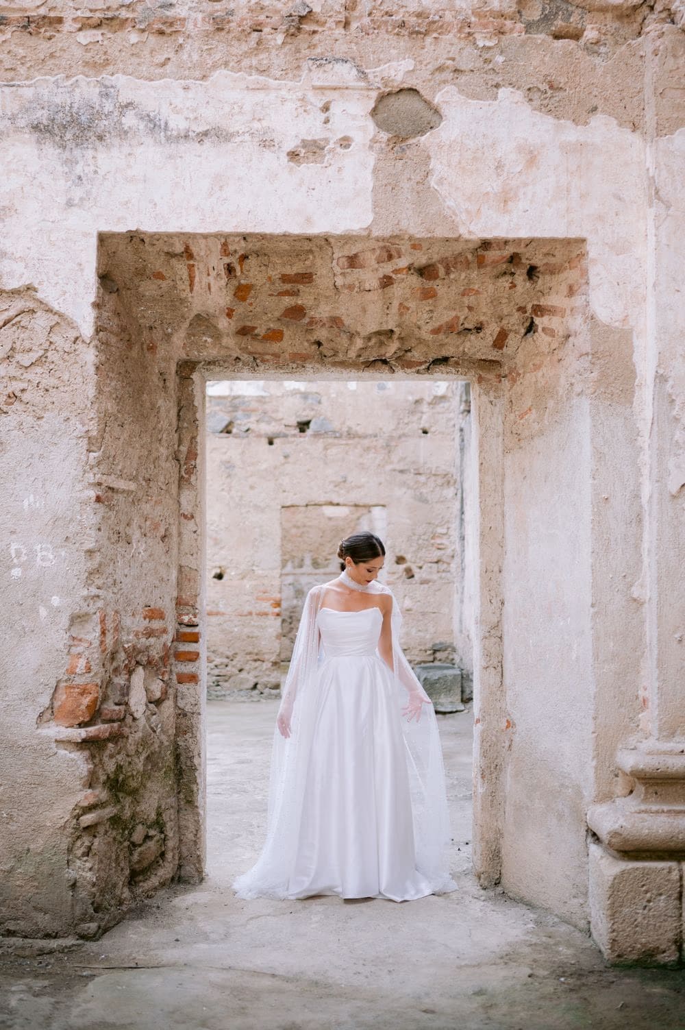 Bride in full ball gown standing under a crumbling stone lintel — quiet ruin portrait in colonial Antigua.