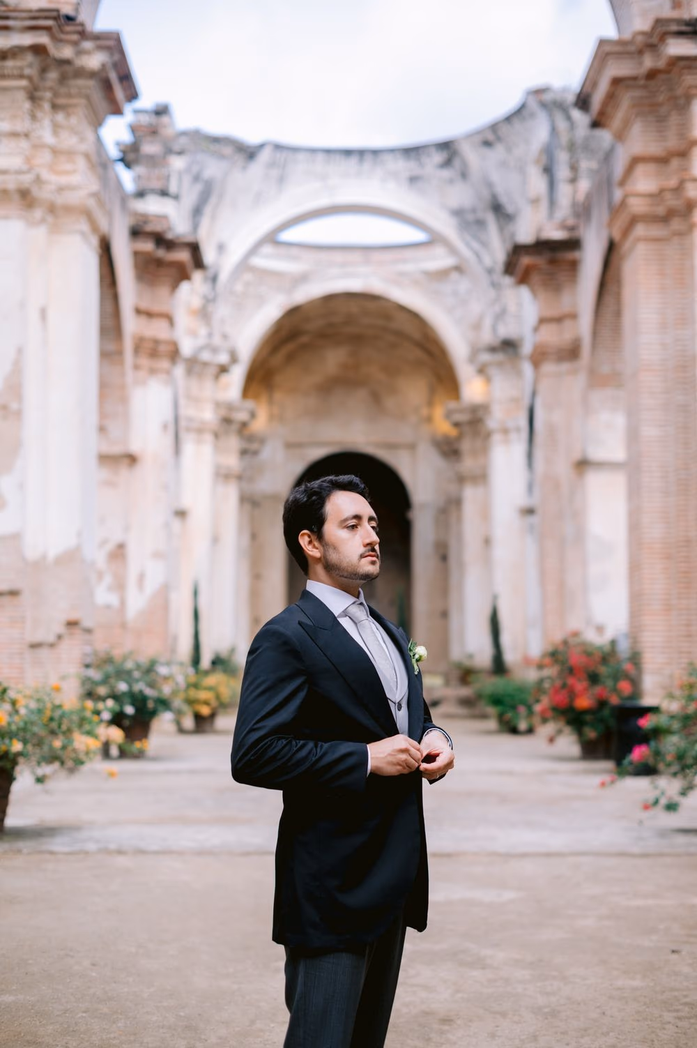 Groom buttons his suit jacket alone in the long ruin arcade flanked by yellow and red flowering planters — Antigua Guatemala portrait.