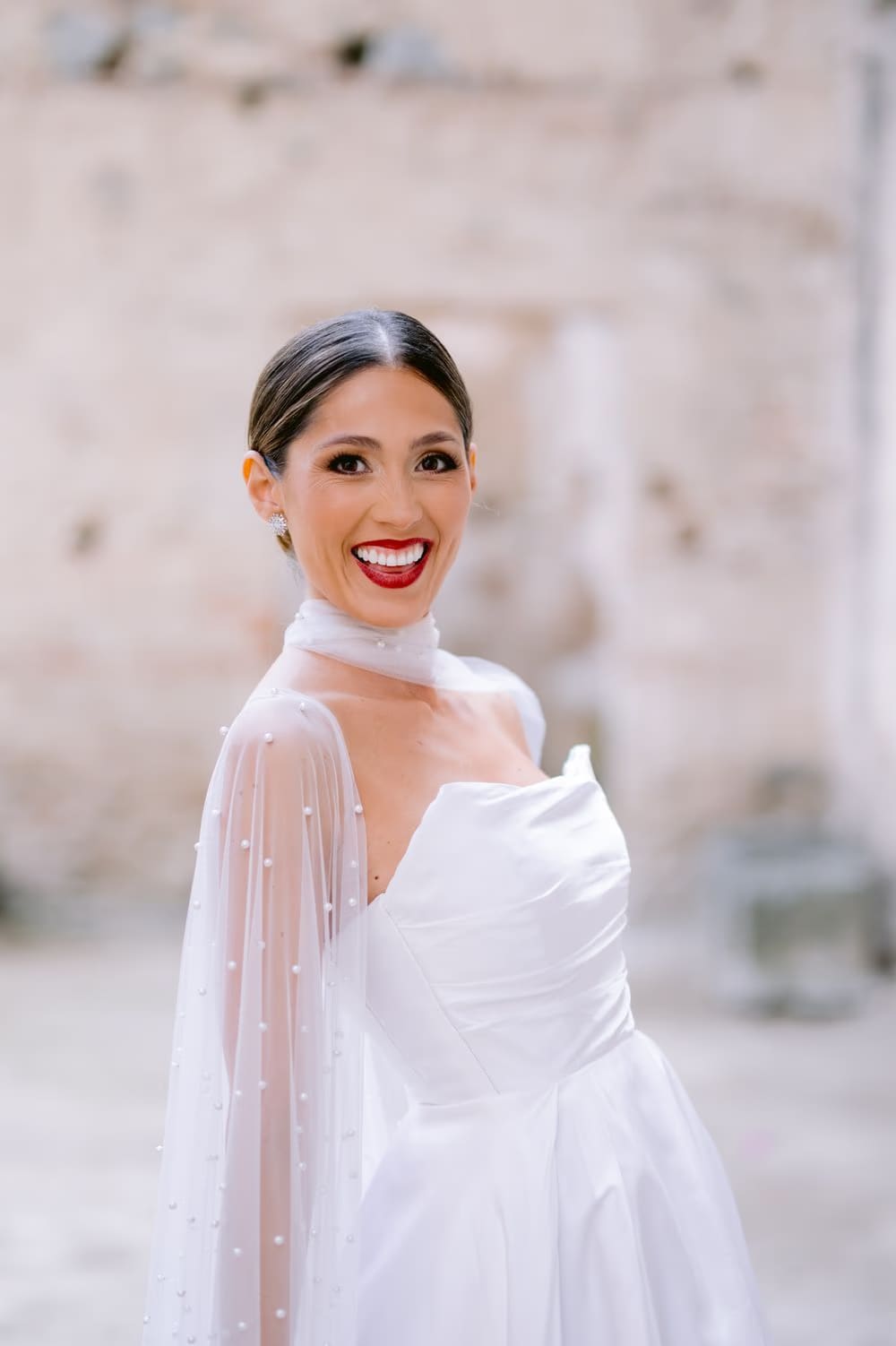 Bride laughs over her shoulder in a candid portrait — pearl-cape sleeves backlit against a soft ruin background.