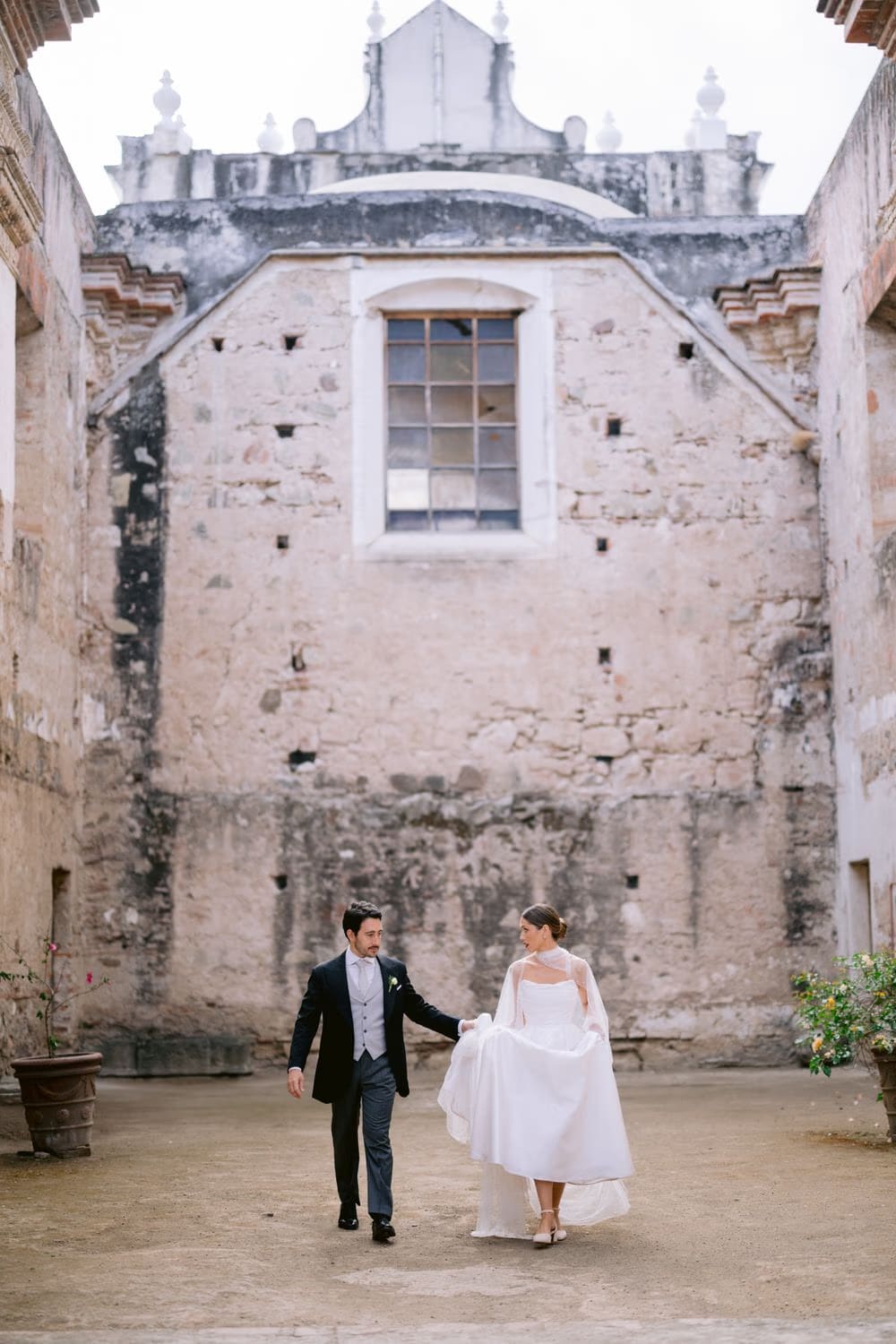Couple walk through a cracked stone courtyard with a tall ruined window behind — destination wedding portrait in Antigua Guatemala.