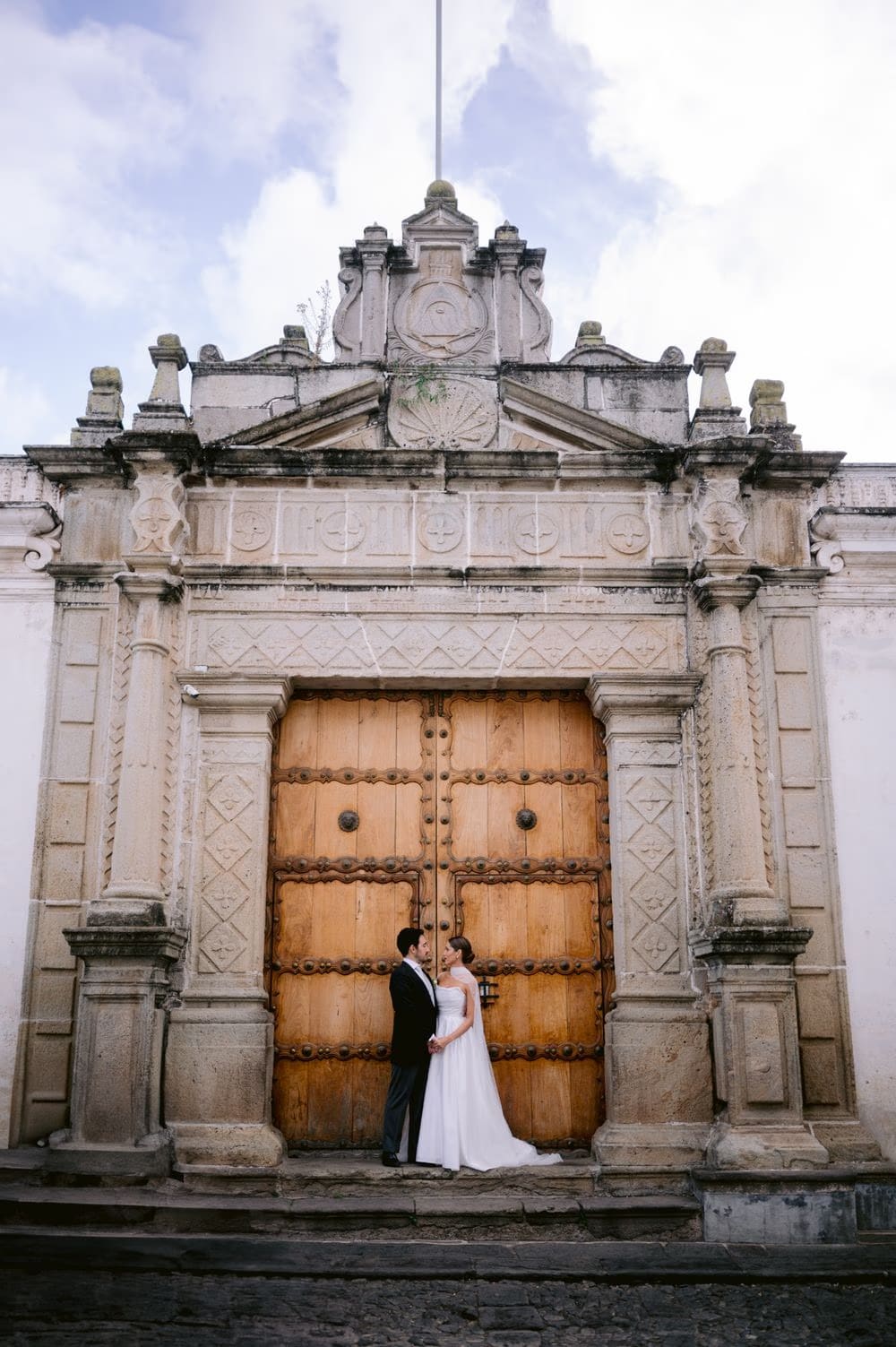 Bride and groom pose in front of a baroque carved wooden door on the cobbled street — colonial Antigua wedding portrait.