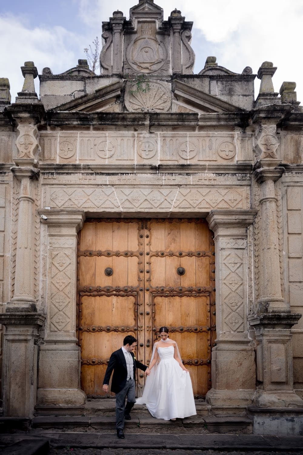 Couple walk down stone steps from a grand carved wooden doorway — wide colonial wedding portrait in La Antigua.