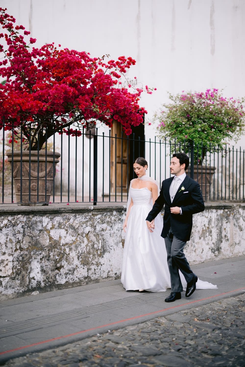 Bride and groom walk a cobbled colonial street past a bougainvillea tree in full red bloom — Antigua wedding portrait.