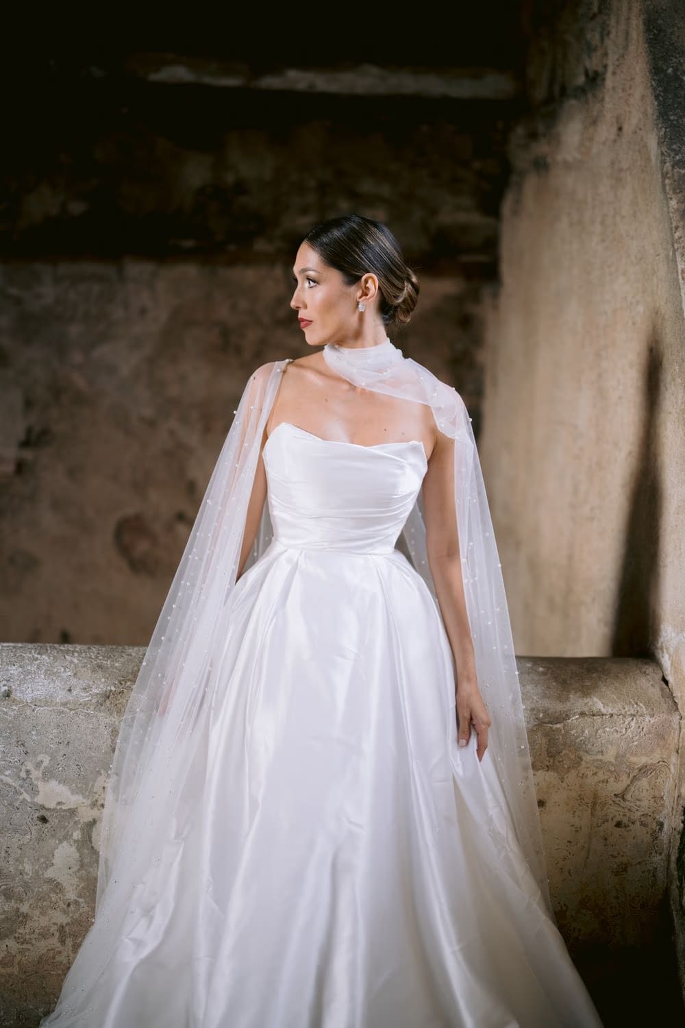 Bride alone in profile inside the stone interior of Capuchinas Ruins — full-length pearl-cape gown editorial portrait.