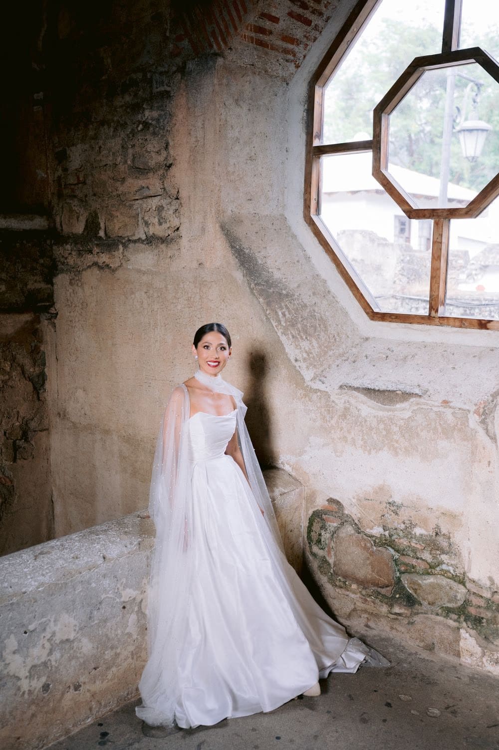 Bride smiles by a large octagonal window in a moss-streaked stone tower — quiet ruin portrait at La Antigua wedding.