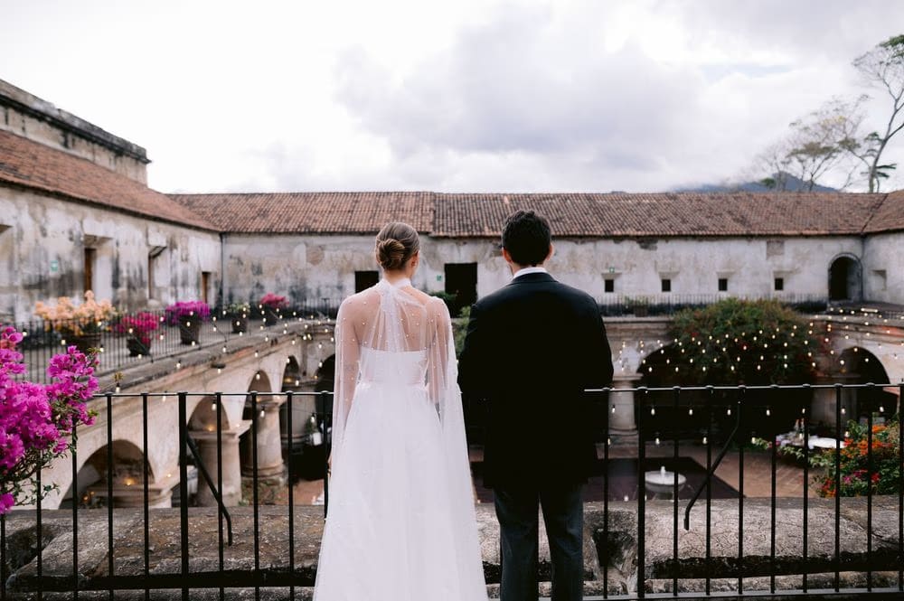 Bride and groom look down from a balcony onto the candle-lit reception courtyard at Capuchinas Ruins as guests begin to arrive.