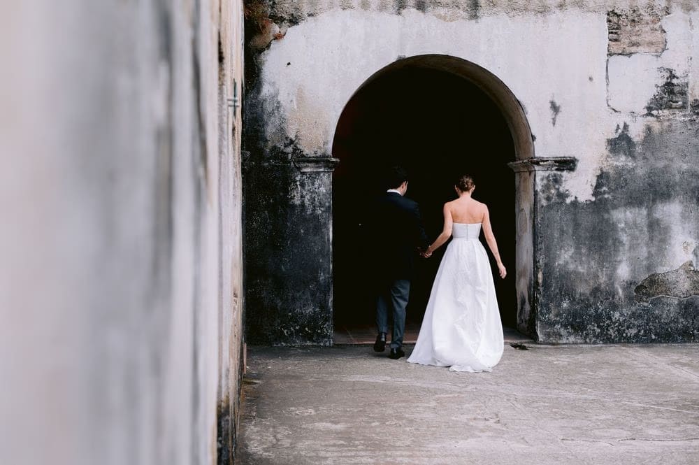Bride and groom walk hand-in-hand through a dark arched passageway between courtyards — Antigua Guatemala wedding moment.