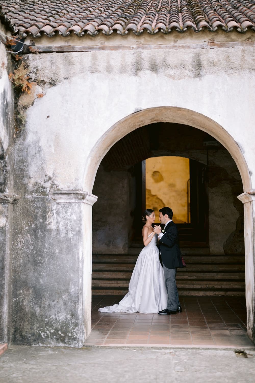 Couple stand close beneath a tile-roofed archway leading into the warmly lit interior — Capuchinas Ruins wedding portrait.