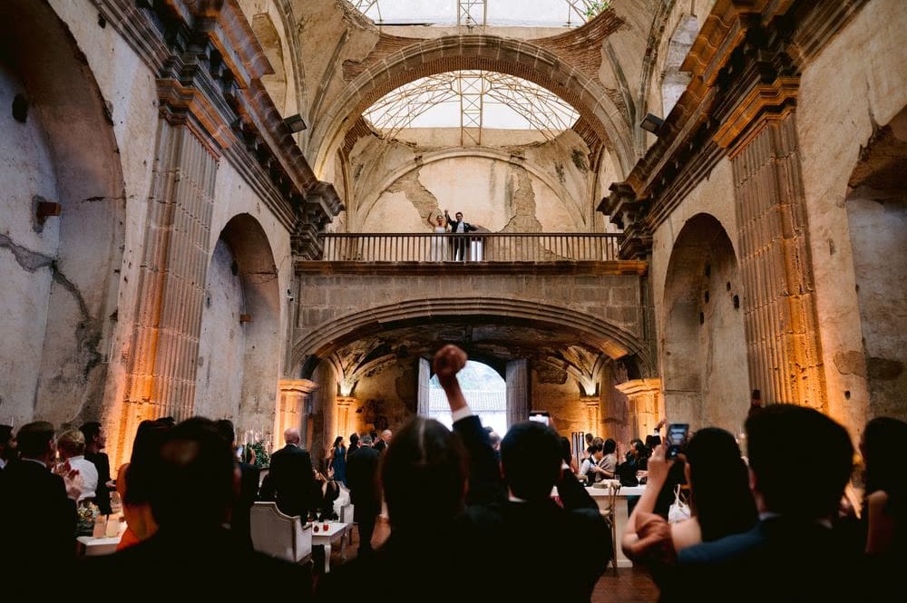 Guests cheer from below as the couple wave from the upper balcony of Capuchinas Ruins — joyful reception entrance.