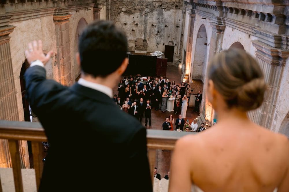 View over the couple's shoulders as they wave to cheering guests below — reception greeting at Capuchinas Ruins.
