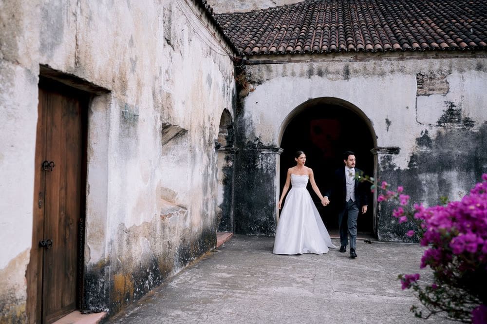 Bride and groom step through an archway by a fuchsia bougainvillea — golden-hour wedding portrait in colonial Antigua.