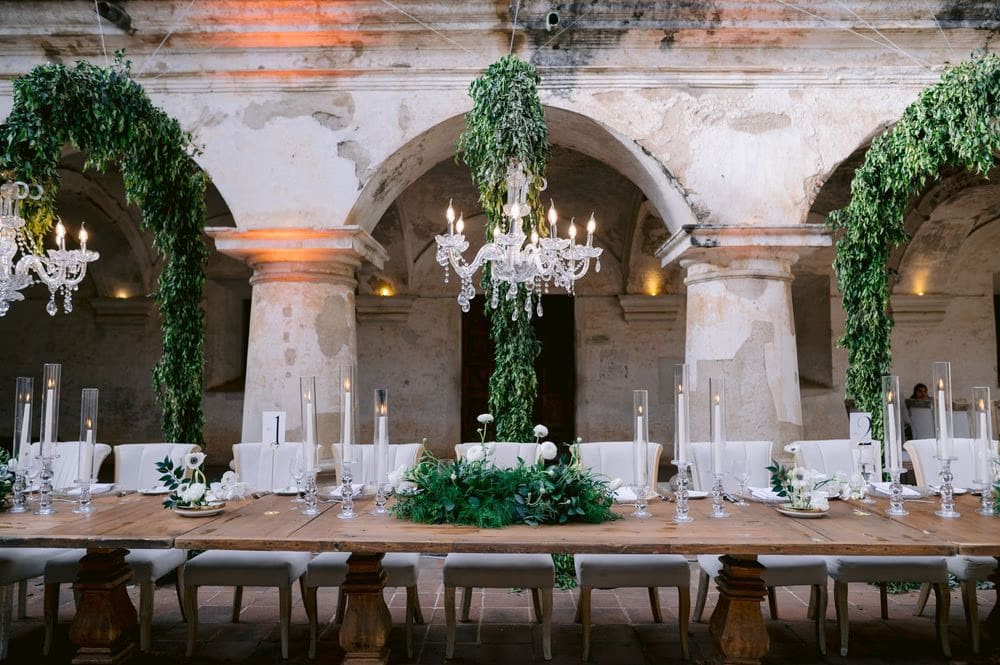 Long banquet table set under hanging greenery and crystal chandeliers in the cloister — candle-lit reception decor.